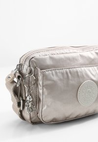 Metallic beige crossbody bag made of soft, textured fabric. Features two zippered compartments, a circular logo patch, and a charm detail.