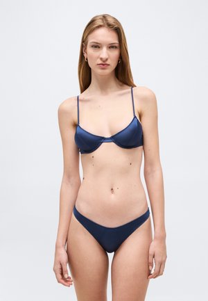 Mladá žena s dlhými blond vlasmi, oblečená v námornícky modrých bikinách, stojaca oproti jednoduchému svetlošedému pozadiu a čelí dopredu.