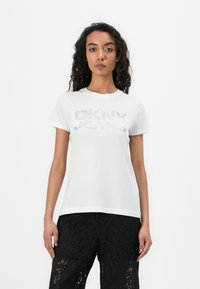 Weißes Baumwoll-T-Shirt mit glänzend silberner Aufschrift "DKNY JEANS". Kurzärmlig und mit Rundhalsausschnitt; kombiniert mit schwarzen Spitzenhosen.