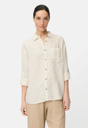 Femme aux cheveux foncés tirés en arrière, portant une chemise beige et blanche à rayures avec des manches retroussées et un pantalon beige.