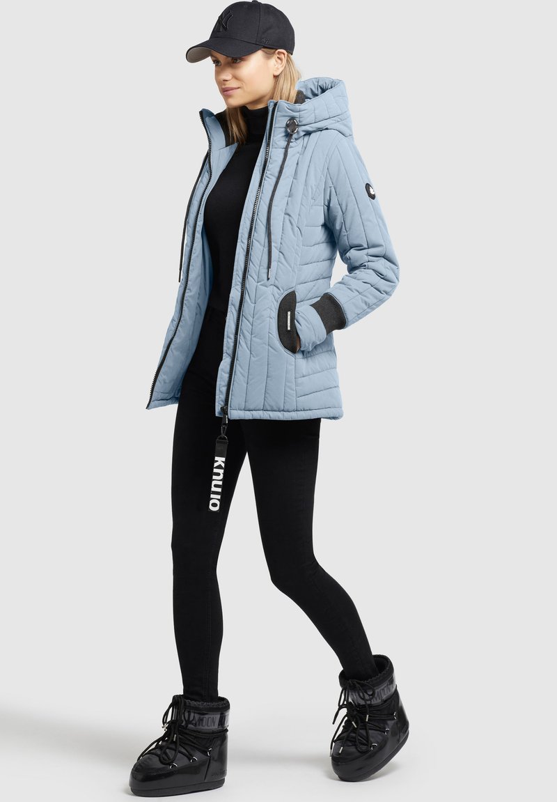 Hellblau gesteppte Jacke mit Kapuze, schwarzen Akzentbündchen, Frontreißverschluss und Kordelzügen. Kombiniert mit schwarzen Leggings und glänzenden schwarzen Stiefeln.