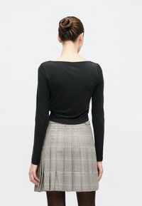 Γυναίκα με καστανά μαλλιά σε κότσο, που φοράει μακρυμάνικη μαύρη μπλούζα και πλισέ γκρι καρό φούστα, στέκεται με την πλάτη προς το μέρος μας μπροστά σε απλό λευκό φόντο.