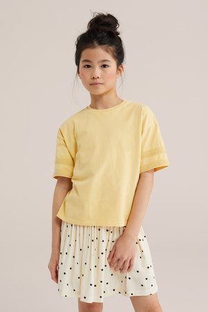 Młoda dziewczyna z ciemnymi włosami związanymi w kok, ubrana w żółty krótki rękaw i białą spódnicę w czarne kropki, stoi na jednolitym tle.