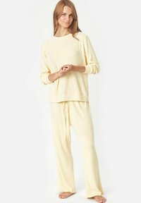 Loungewear komplektas švelnaus geltonos spalvos grįžto audinio. Sudėtyje yra ilgomis rankovėmis marškiniai su apvalia iškirpte ir laisvai krentančios kelnių su raišteliu. Lygi tekstūra.