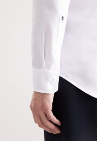 Camicia bianca con motivo a spina di pesce; presenta un polsino con bottone e un piccolo dettaglio ricamato di rosa sulla manica.