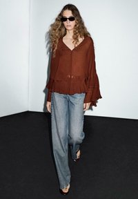 Top marrone a maniche lunghe con orlo a frange e scollo a V profondo, abbinato a jeans a gamba larga di colore blu chiaro. Occhiali da sole neri completano l'outfit.