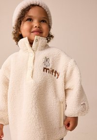 Valge fliispusa kõrge kaelusega, millel on nööbiga kinnis ja kirjaga "miffy" logo. Pehme tekstuur ja mugav disain, külgpocketitega.