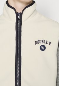 Chaqueta de forro polar en crema y gris con cuello alto, cremallera frontal y logotipo bordado "DOUBLE A" en el pecho. Textura suave.