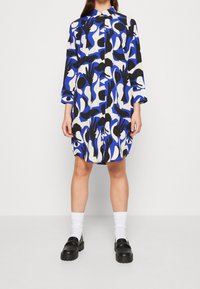 Šaty košilového typu s dlouhými rukávy v modré, černé a bílé abstraktní šabloně, s knoflíkovou légou a límečkem. V kombinaci s černými botami a bílými ponožkami.