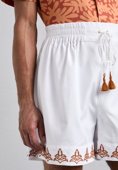 White shorts with a textured finish, featuring an elastic waistband and drawstring, accented with intricate orange embroidery at the hem.