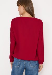 Rød langærmet bluse med en bred halsudskæring, der har en glat tekstur og en afslappet pasform. Båret med blå denim jeans med synlige baglommer.