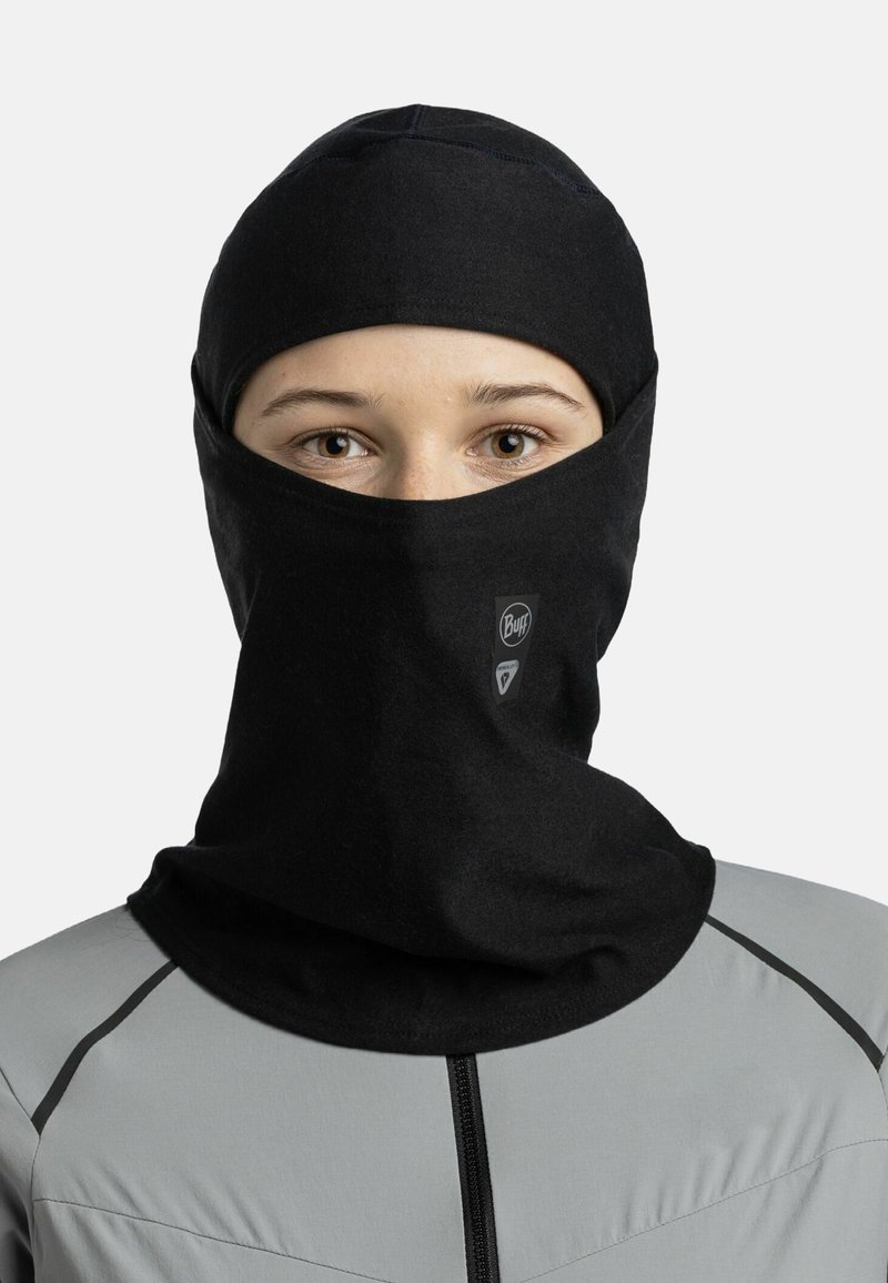 Black balaclava made of soft fabric, featuring a snug fit and a logo on the front. Worn with a gray, fitted jacket with subtle seams.