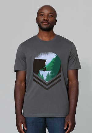 Man draagt een grijs shirt met een afbeelding van een rotsklimmer die aan een klif hangt, met bomen en bergen op de achtergrond.