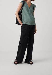 Grön texturerad v-ringad topp med volangärmar, parat med lösa svarta byxor. Skorna är svarta sandaler med dekorativt spänne.