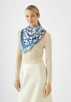 Vrouw met blond haar in een staart draagt een beige trui, een witte rok en een blauwgepatteerde sjaal om haar nek tegen een effen achtergrond.