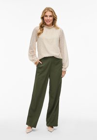 Ljus beige stickad blus med vågmönster och puffärmar, ihop med olivgröna vida byxor. Tan spetsiga skor fulländar looken.
