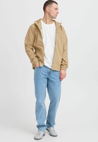 Light brown hooded jacket with elastic cuffs, worn over a white t-shirt and blue jeans, paired with gray sneakers.
