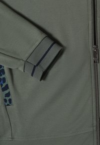 Manche de veste verte avec cuff côtelé présentant deux bandes foncées, à côté d'une fermeture éclair argentée et d'une poche avec un bord motif camouflage.