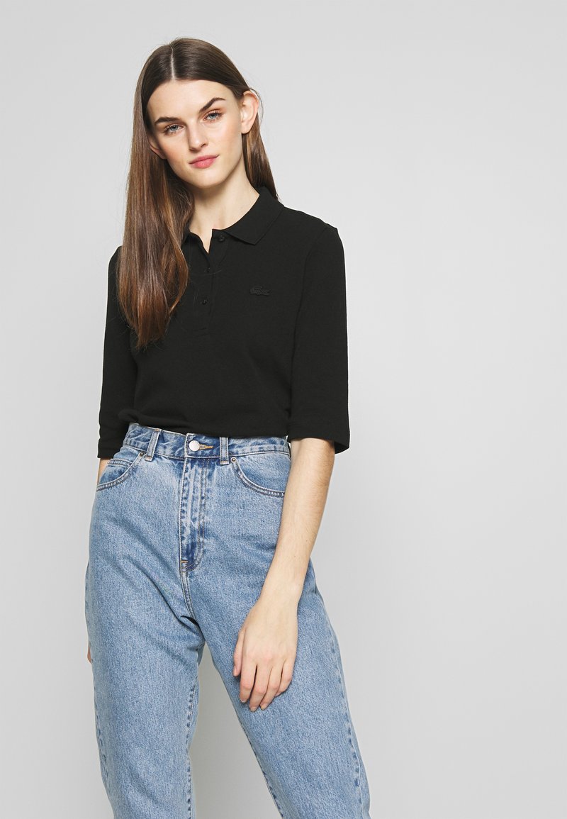 Svart polo-skjorte med trekvart ermer og krage, kombinert med høyhalsede lyseblå denim jeans. Lacoste-logo i svart på skjorten.