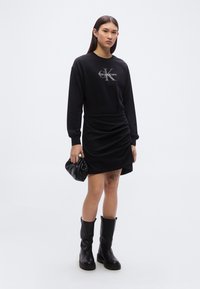 Μαύρο φούτερ με κουκούλα και λογότυπο Calvin Klein, σε συνδυασμό με μαύρη φούστα με ρεβέρ και μαύρες μπότες μέχρι το γόνατο. Το μοντέλο κρατά μια μικρή μαύρη τσάντα.