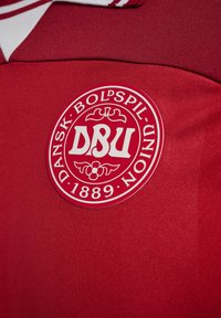 Röd sporttröja med en rund logotyp med "DBU" i mitten, omgiven av "DANSK BOLDSPIL-UNION" och "1889" längst ner.