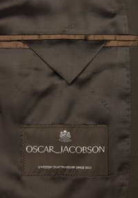 Brun tyginteriör med fiskbensmönster, med en etikett med "OSCAR_JACOBSON" och en logotyp, accentuerad med ett ljusbrunt band.