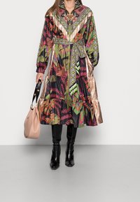 Abito floreale colorato con colletto, gonna a strati e dettagli metallici; abbinato a stivali neri alti e una borsa beige.