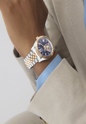 Uređaj od nehrđajućeg čelika i ružičastog zlata s tamnoplavim brojčanikom, rimskim brojevima i prozirnim kućištem, nošen na teksturiranom narukvici.