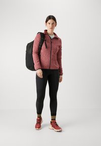 Punainen marle-kuvioinen vetoketjullinen fleece-takki, mustat legginsit taskuilla, punaiset urheilukengät ja musta reppu. Yksinkertainen muotoilu, jossa on toiminnallisia ominaisuuksia.