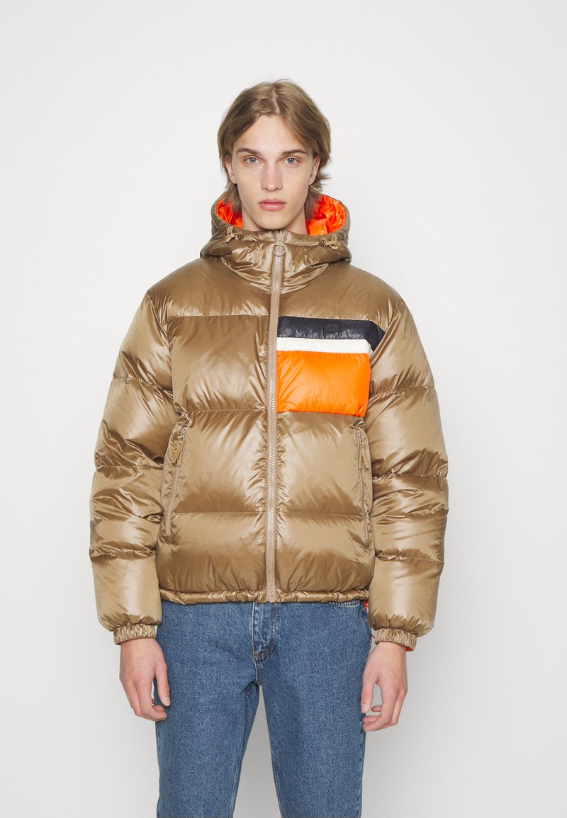 Braune gepolsterte Jacke mit Kapuze, ausgestattet mit einer orangefarbenen Brusttasche, schwarzen und weißen Akzenten sowie elastischen Bündchen; glatte, glänzende Textur.