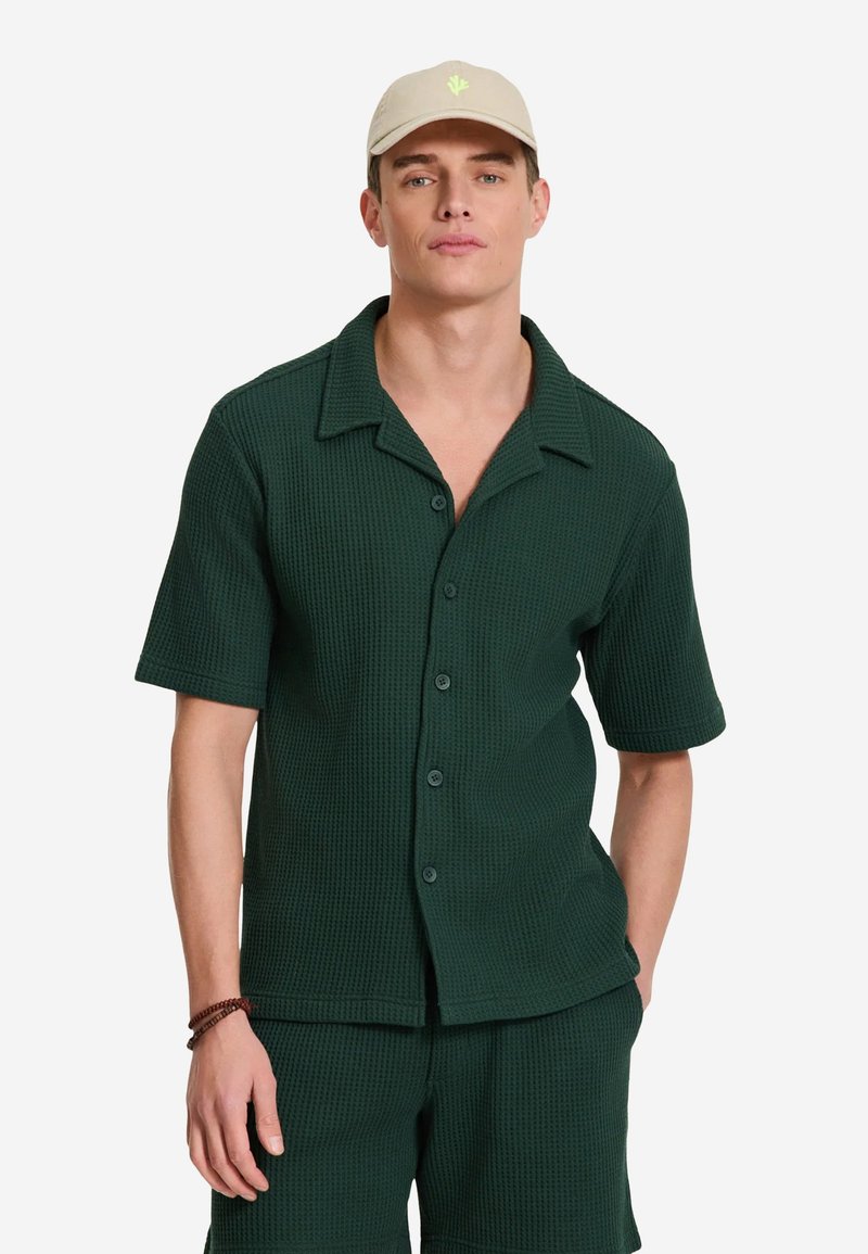 Mand iført beige baseballkasket, mørkegrøn kortærmet skjorte med knapper og matchende shorts, stående foran en ensfarvet hvid baggrund.
