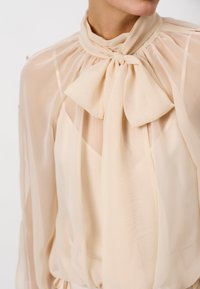 Beige Seidenbluse mit hohem Kragen und großer Schleife. Durchsichtiger Stoff mit fließenden Ärmeln und leicht lockerer Passform.