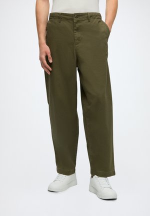Homme portant un pantalon ample vert olive et des baskets blanches, debout devant un fond uni, une main dans la poche.