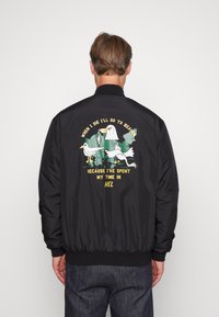 Giacca bomber nera con colletto e polsini a coste. Sul retro presenta un design grafico colorato di uccelli e testo in giallo e bianco.