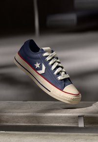 Zapatilla baja azul con cordones blancos y logo de estrella, suela beige con franjas rojas y azules, flotando sobre un banco de piedra.