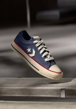 Blue low-top sneaker with white laces and star logo, beige sole with red and blue stripes, hovering above a stone bench.
