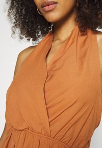 Šaty barvy rezavé s halter výstřihem, vyrobené z měkké látky, s nařaseným pasem a splývavým živůtkem pro uvolněnou siluetu.