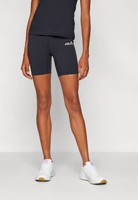 Črni elastični kolesarski kratki hlače z gladko teksturo in belim logotipom. Nošene s črnim oprijetim topom in belimi športnimi čevlji.