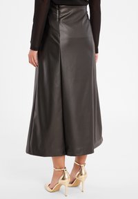 Schwarzer Kunstleder A-Linien-Maxi-Rock mit glatter Textur, seitlichem Reißverschluss und fließender Silhouette. Kombiniert mit goldenen Riemchen-Absätzen.