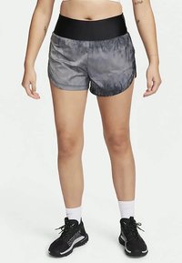 Pernas de uma pessoa a usar calções de corrida em tie-dye preto e cinza, meias brancas até ao tornozelo e sapatos de corrida de trilha pretos em Gore-Tex, de pé sobre um fundo branco.