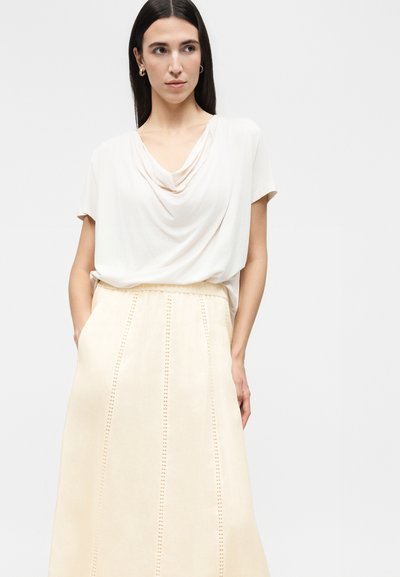 Γυναίκα με μακριά σκουρα μαλλιά που φοράει χρυσούς κρίκους σκουλαρίκια, λευκό κοντομάνικο τοπ με ρίγες και φούστα εκρού με ψηλή μέση και κάθετες λεπτομέρειες.