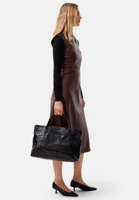 Borsa tote in pelle nera con embossing a forma di coccodrillo, caratterizzata da doppini manici, forma strutturata e zip in alto. Indossata con un vestito in pelle marrone.
