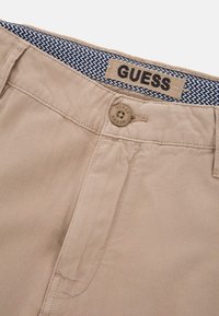 Chinos in cotone beige con chiusura a bottone, caratterizzati da un'etichetta "GUESS" sulla vita e un rivestimento interno a motivo chevron blu navy.