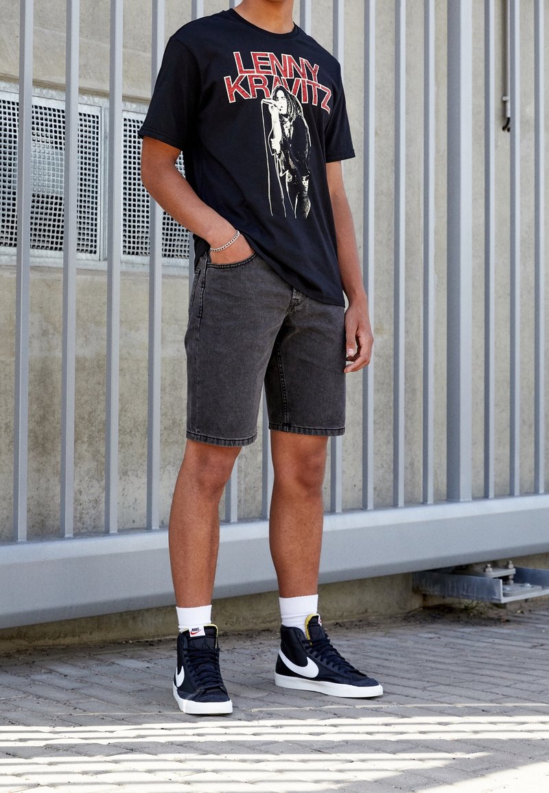 Persona con una camiseta negra de Lenny Kravitz, pantalones cortos de mezclilla gris oscuro, calcetines blancos y zapatillas altas negras de Nike de pie al aire libre.