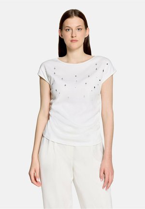 Femme portant un haut blanc à manches courtes avec de petites décorations métalliques dispersées et un pantalon blanc ample, debout devant un fond uni.