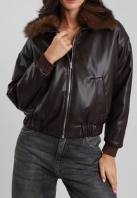 Giacca bomber in pelle marrone con colletto in pelliccia sintetica, chiusura con zip, polsini a coste e vita elastica. Presenta una tasca frontale.