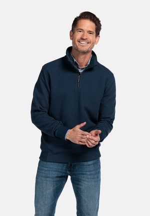 Donkerblauwe ritspull met een gestructureerde stof, hoge kraag en geribbelde manchetten, gecombineerd met een geruit overhemd eronder en denim spijkerbroek.