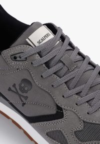 Zapatilla gris con materiales de tejido y ante, acentos texturizados, un logo de calavera y cordones planos. Suela de goma con detalles en blanco y marrón.