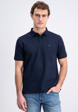 Homme aux cheveux bouclés portant un polo bleu foncé à manches courtes et un jean bleu clair, debout avec une main dans la poche.