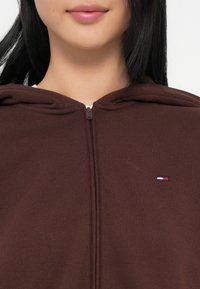 Brown zip-up hoodie made of soft fabric, featuring a hood and a small logo in red, white, and blue on the left chest area.